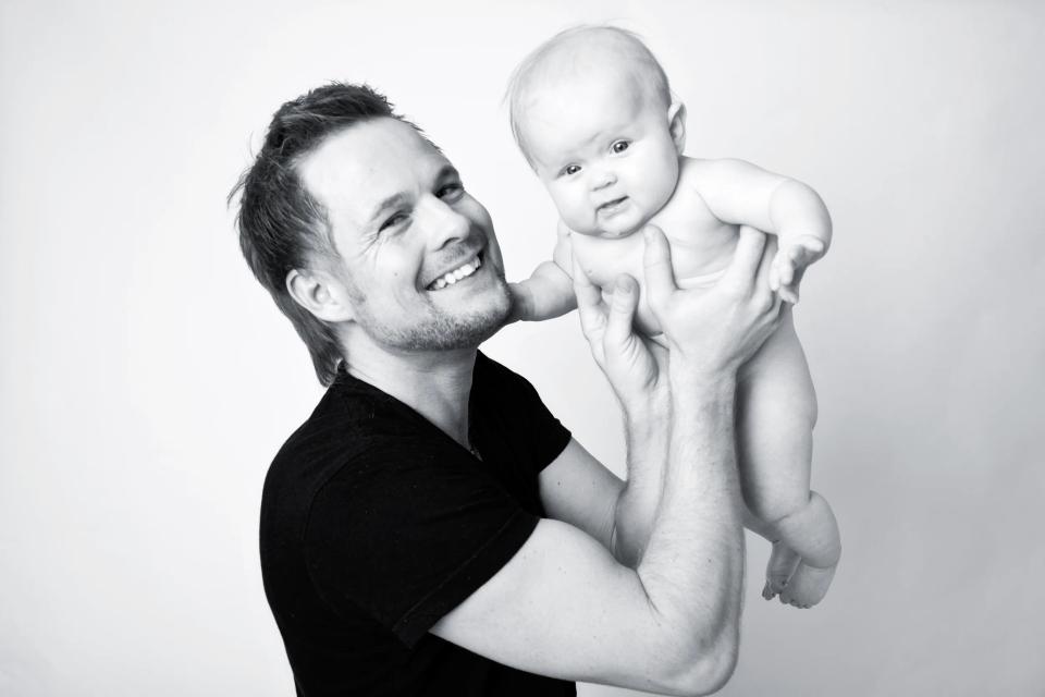 Sort-hvidt babyfoto hvor en smilende far holder sin baby op i luften med blødt, naturligt lys. Stemning af glæde.