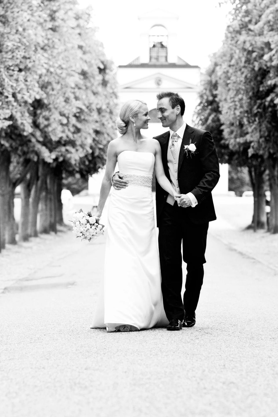 Bryllupsfotografering af brudepar, der går hånd i hånd og smiler forelsket mod hinanden foran Hørsholm Kirke.