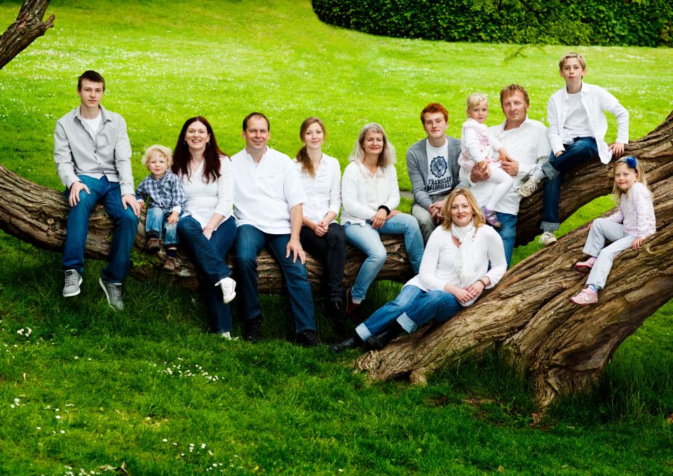 Familiefotografering: Stor familie sidder smilende på en tyk træstamme i grønt græs under åben himmel.