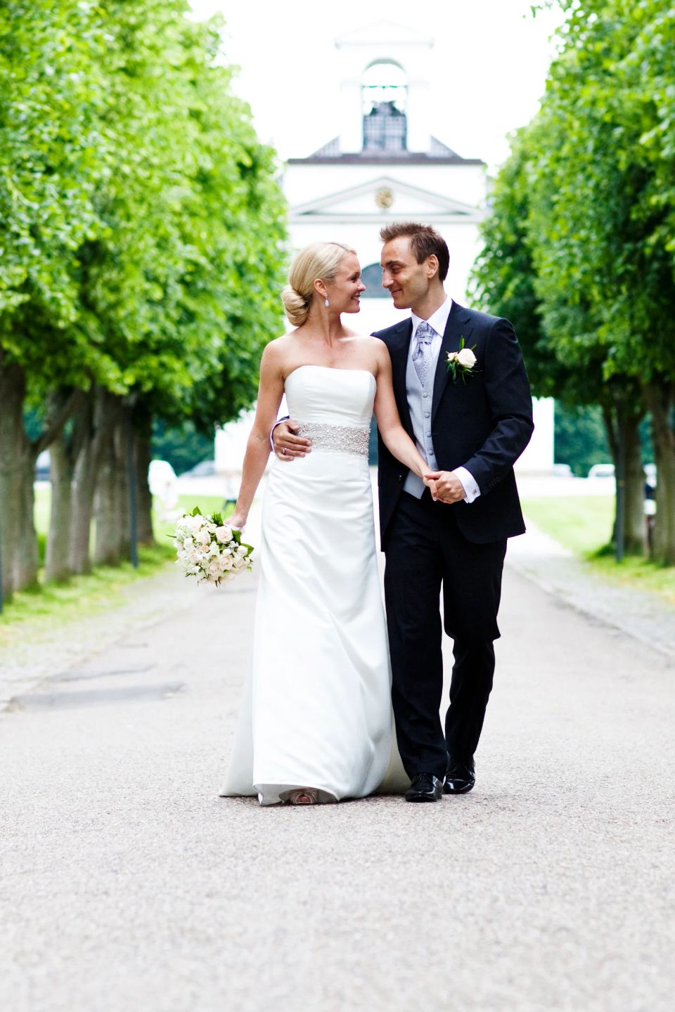 Bryllupsfotografi af brudepar, der smiler og holder i hånd på allé med grønne træer og kirke i baggrunden.