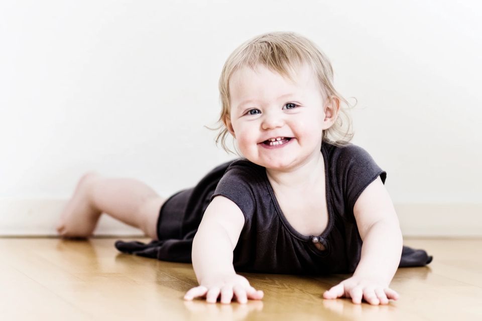 Glad baby kravler på trægulv med stort smil. Baby fotografering i lyse farver med naturligt lys.