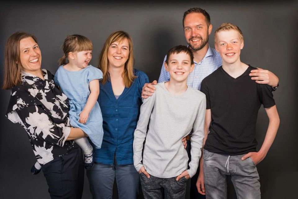 Familiefoto af stor, glad familie med forældre, en pige og tre drenge, smilende mod kameraet på mørk baggrund.