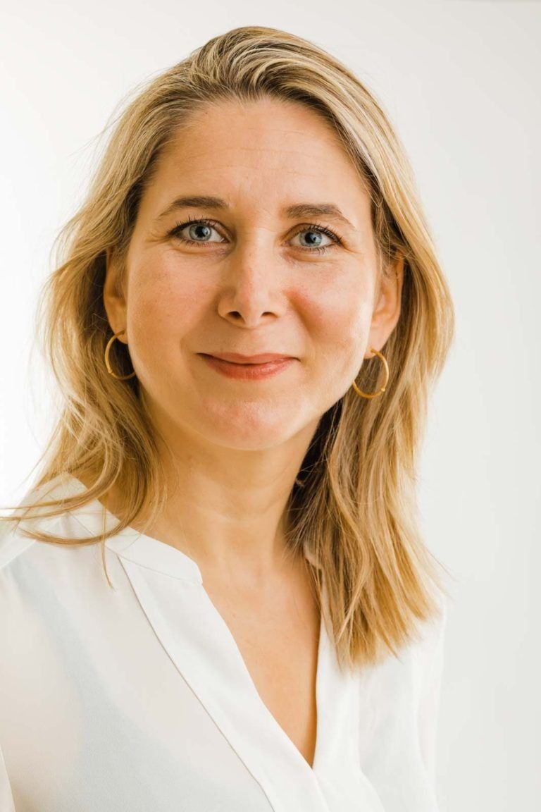 Portrætfoto af en blond kvinde med et roligt, autentisk smil mod en lys, minimalistisk baggrund, iført en hvid skjorte.