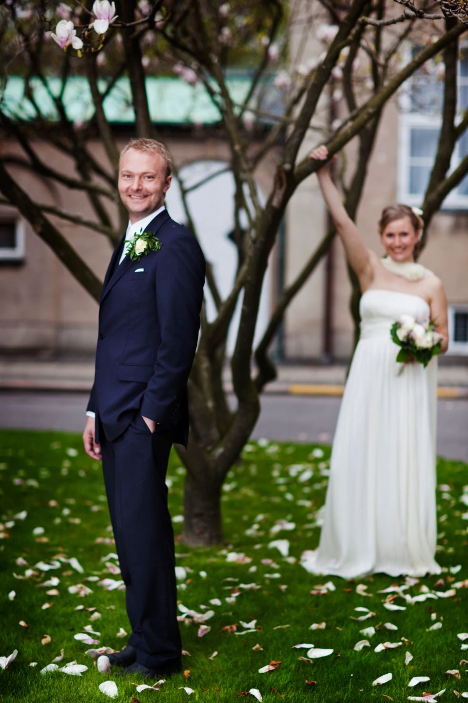 Bryllupsfoto af smilende brudepar udendørs med lyserødt magnolietræ og spredte kronblade på græsset i naturligt lys.