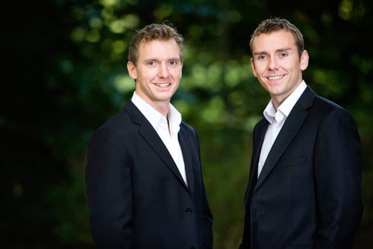 Portrætfoto af to smilende mænd i sorte jakkesæt med hvide skjorter. De står udenfor med sløret grøn baggrund.