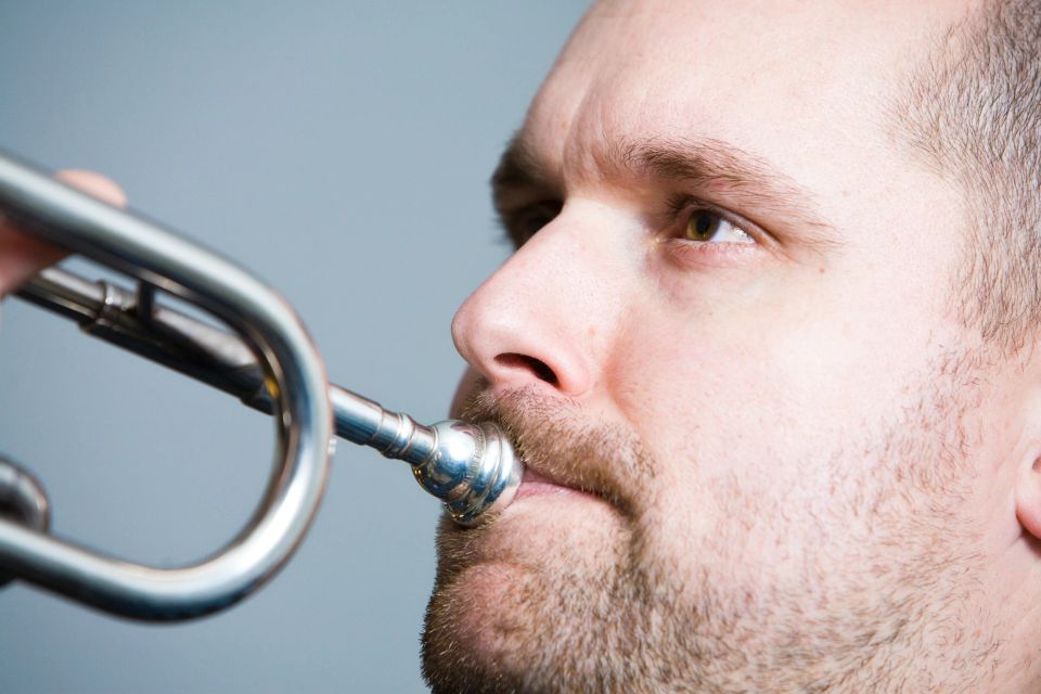 Nærbillede af mands ansigt der spiller på trompet; man ser mundstykket mod hans læber. Portrætfoto.