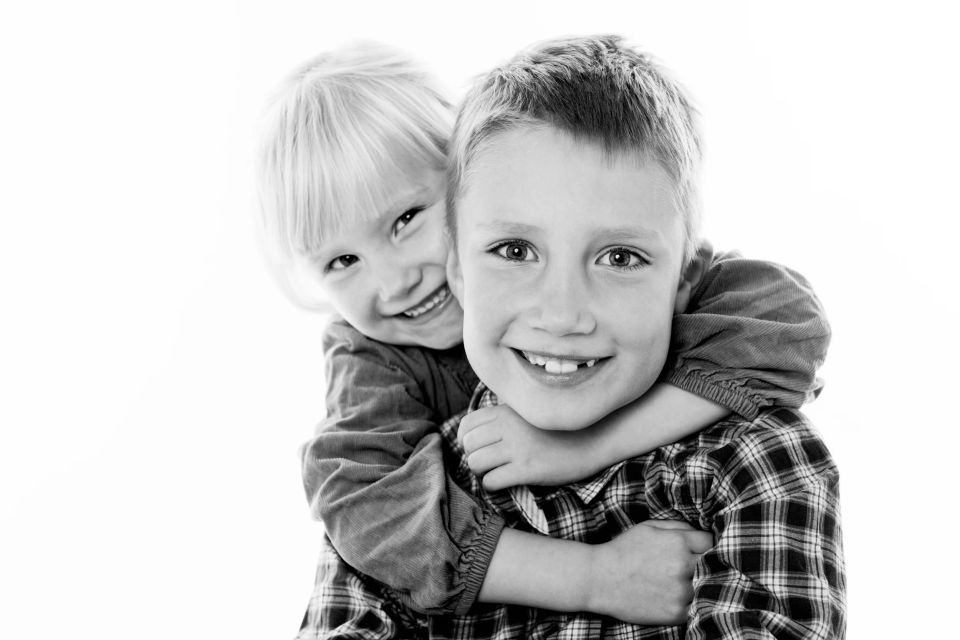Sort-hvid børnefoto af to glade søskende. Den ældre dreng smiler til kameraet, imens lillesøster omfavner ham bagfra.
