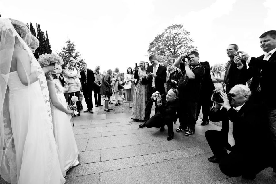Bryllupsfotografering af to brude foran en flok gæster, der begejstret tager billeder. Sort/hvid, naturligt lys.