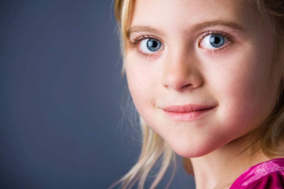 Børnefotografering: Nærbillede af piges ansigt med store blå øjne, lyst hår og fin belysning. En glad barn. 