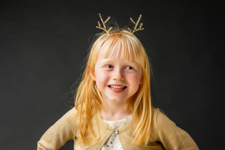 Børnefoto af smilende, blond pige med gyldent gevir-hårbånd og gul cardigan mod mørk baggrund.