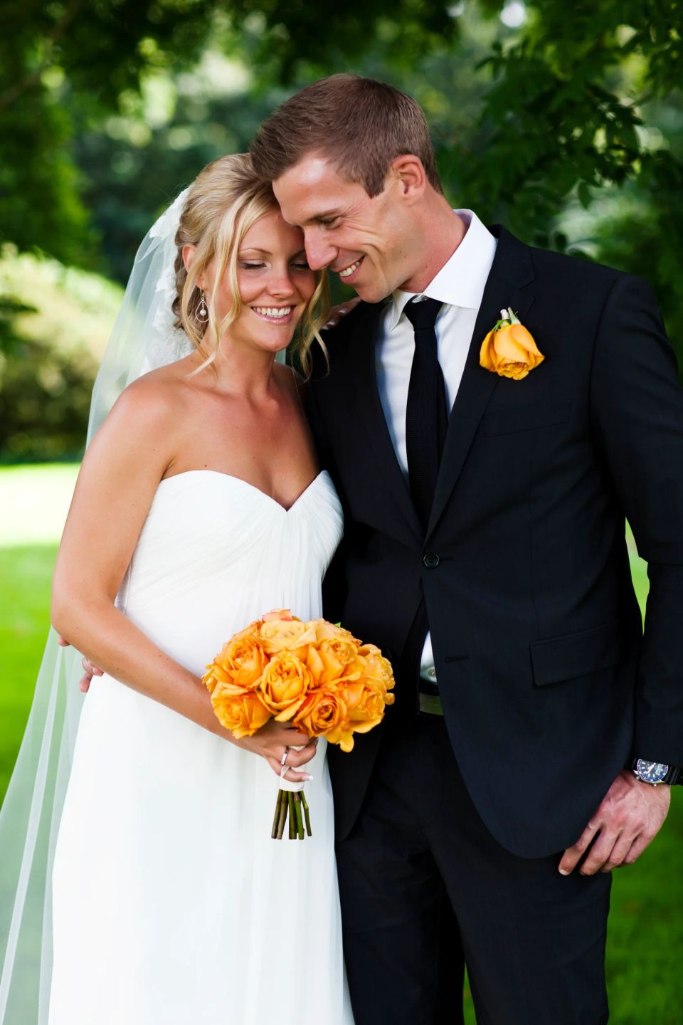 Bryllupspar, der omfavner hinanden i grønne naturlige omgivelser, med brudebuket af orange roser.