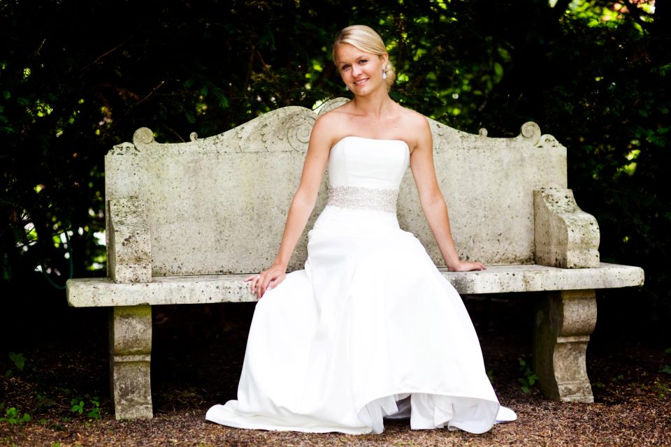 Bryllupsfotografering af smilende brud i hvid kjole med rhinstensbælte, siddende på stenbænk i grønne omgivelser.