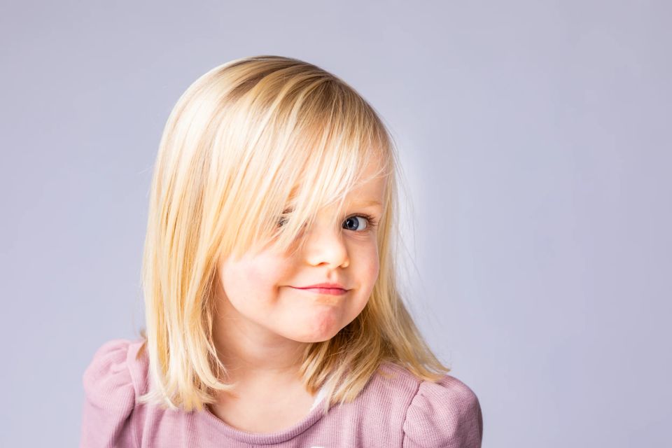 Naturligt portrætfoto af lysthåret barn i lyserød bluse. Legende blik, håret dækker let det ene øje.
