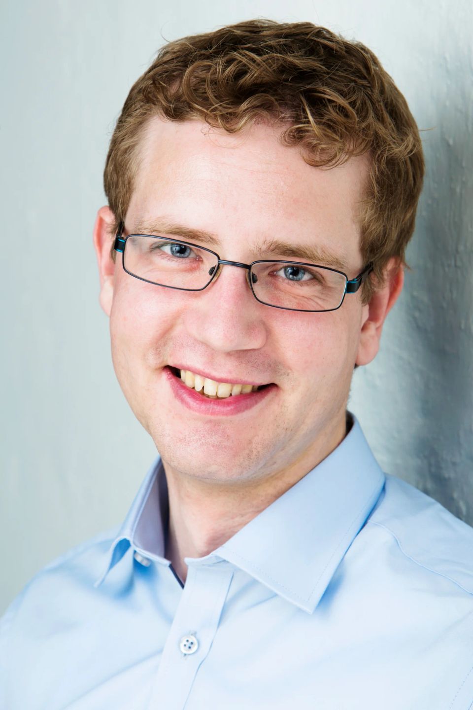 Nært portrætfoto af smilende mand med lyst, krøllet hår, blåt blik og briller, iført lyseblå skjorte.