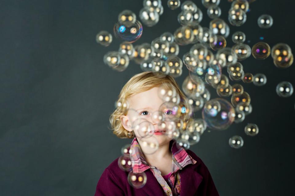 Børnefoto af smilende, blond pige der kigger nysgerrigt frem bag mange flyvende sæbebobler mod mørk baggrund.