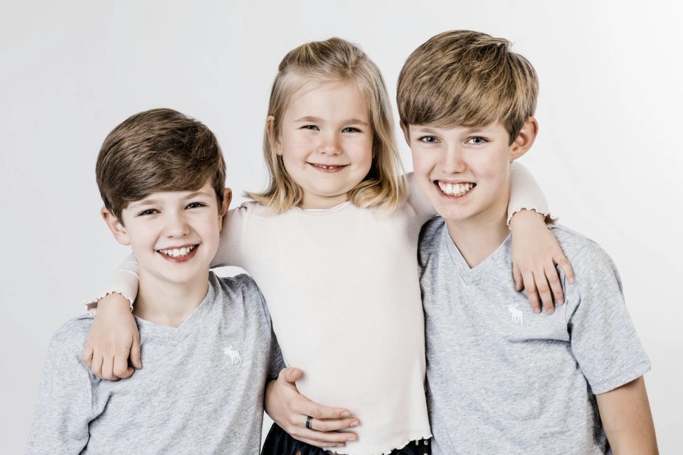 Tre glade børn i en børnefotografering, der smiler ind i kameraet med armene om hinanden på en hvid baggrund.