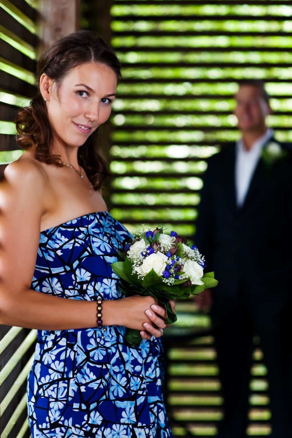 Glad bryllupsgæst i blå kjole med farverig blomsterbuket, en mand ses sløret bagved i naturligt lys.