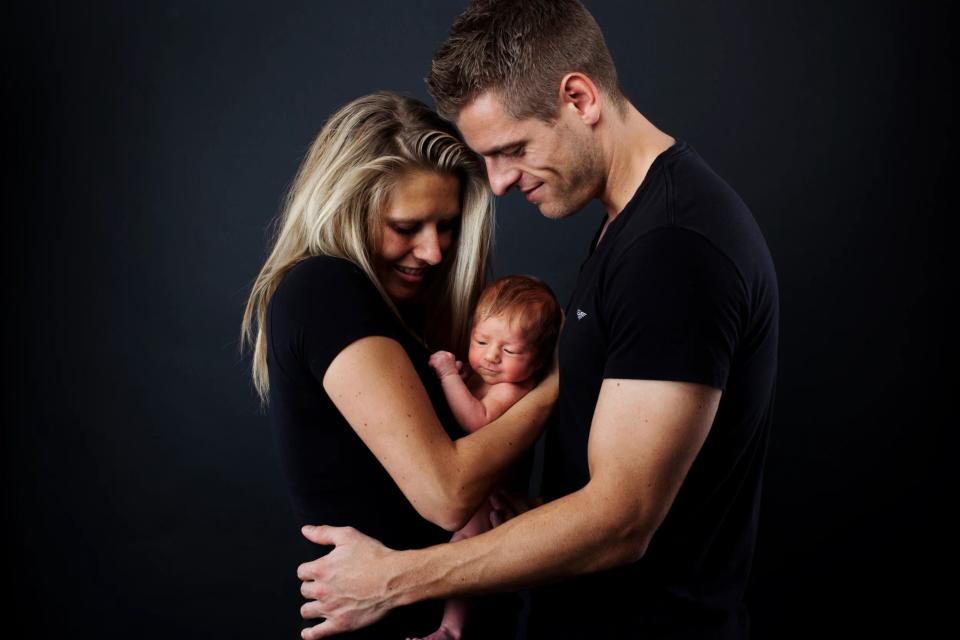 Familiefoto af forældre, der ømt holder deres nyfødte baby foran en mørk baggrund i blødt sidelys.