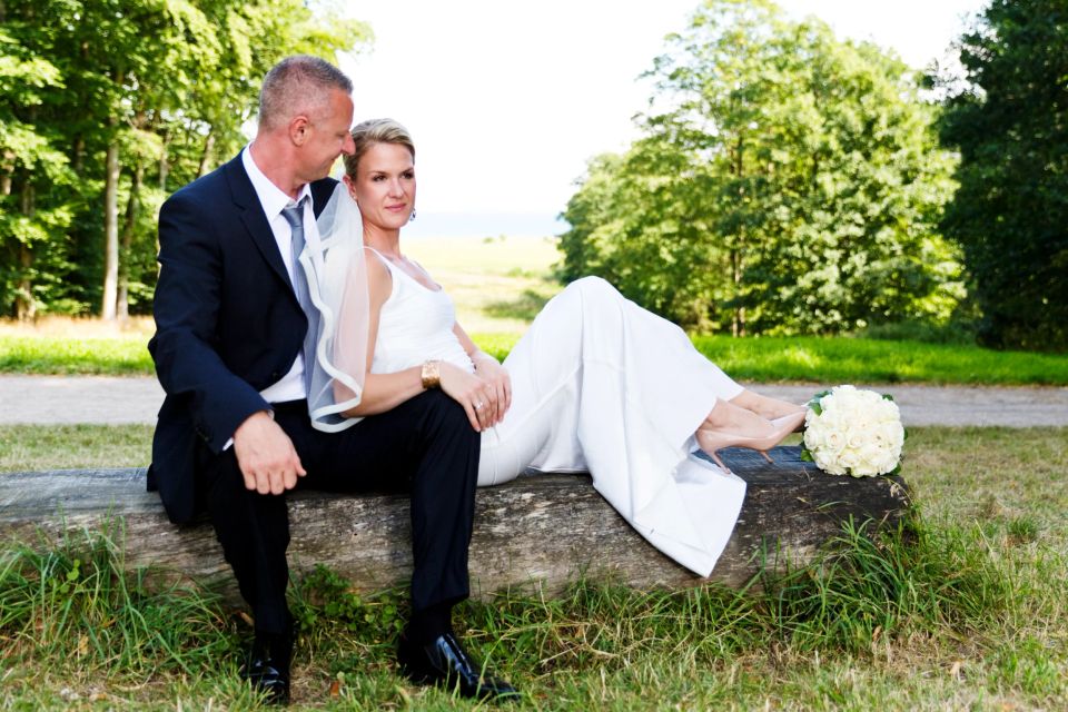 Bryllupsbillede af par siddende på træstamme, bruden i hvidt, brudgommen i mørkt jakkesæt. Natur baggrund og rolig stemning.