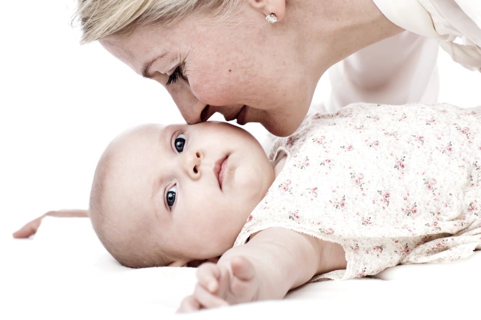 Nærbillede af babyfotografi, hvor en mor kysser sin baby på panden. Barnet ligger ned og kigger direkte mod kameraet.