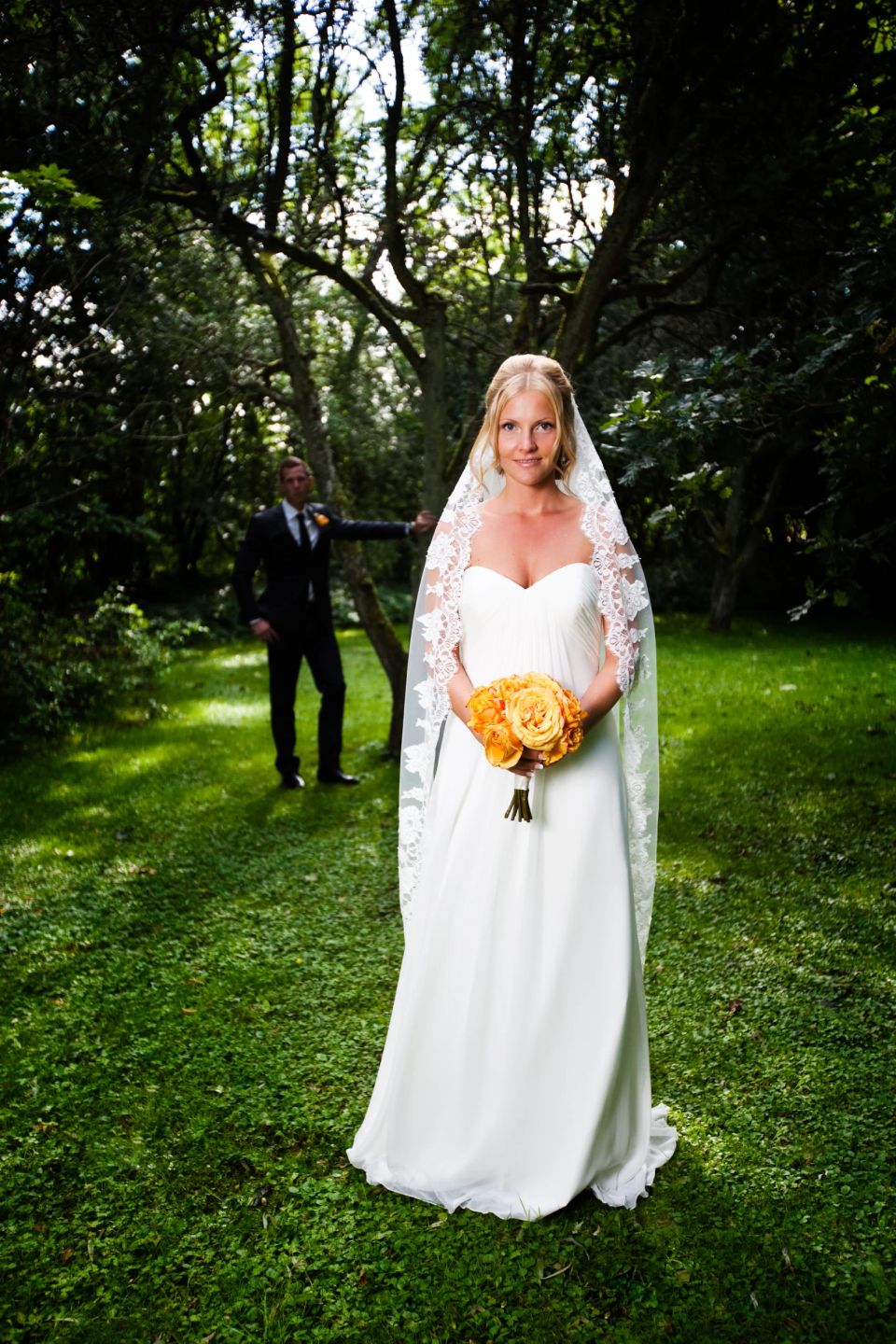 Brudeparrets første blik i grøn natur. Bruden smiler, mens gommen venter i baggrunden bag et træ. Bryllupsfoto.