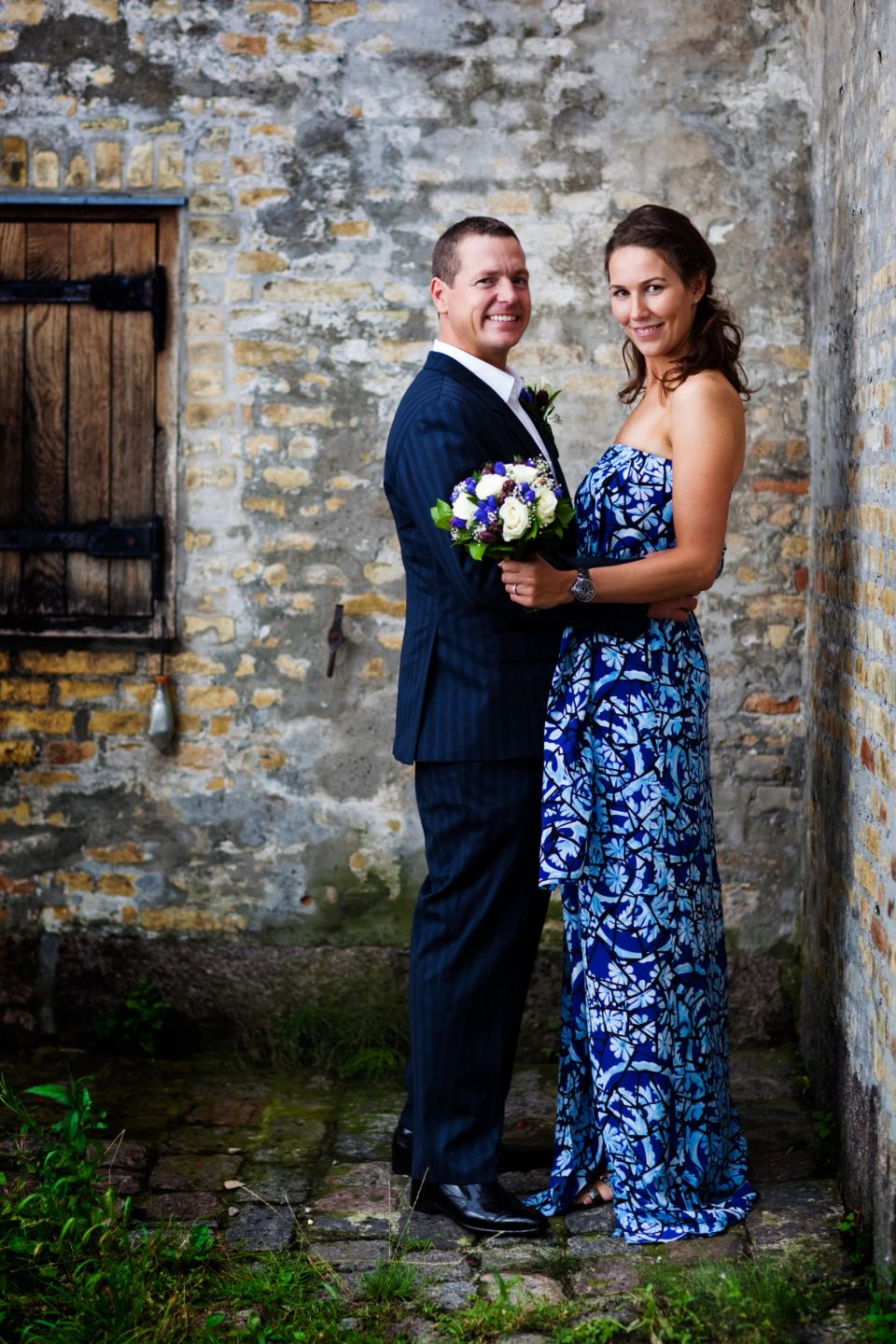 Bryllupsfotografi: Et par smiler varmt til kameraet foran en rustik murstensvæg, kvinden med en brudebuket i hånden.