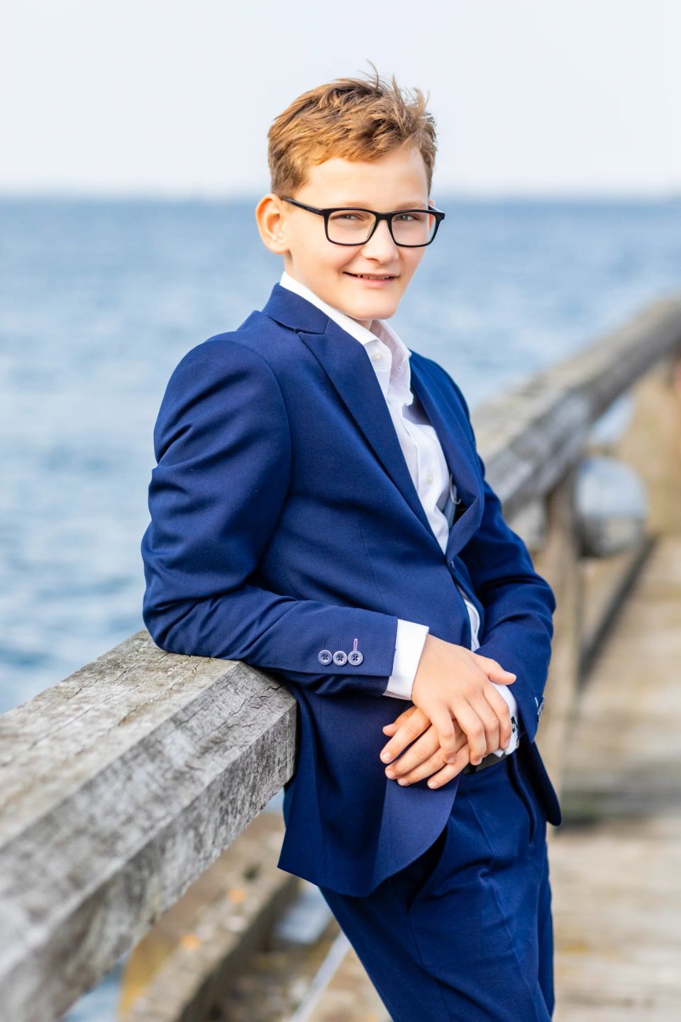 Portrætfoto af ung konfirmand i blåt jakkesæt, smilende mod kameraet, med havets blå nuancer i baggrunden.