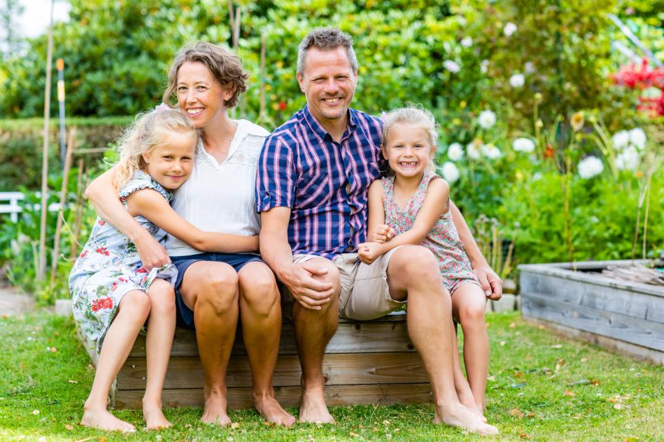 Familiefotografering: Smilende familie med to piger sidder barfodet på en træbænk i haven i blødt dagslys.