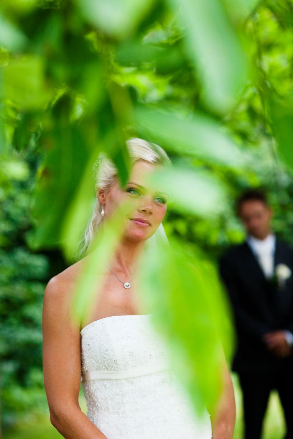 Bryllupsfotografering med smuk brud i hvid kjole og elegant halskæde, indrammet af grønne blade i forgrunden.