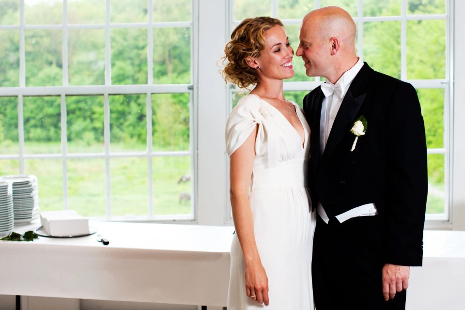 Bryllupsfotografering: Smilende brudepar i naturligt lys foran et stort vindue med grønne træer udenfor.