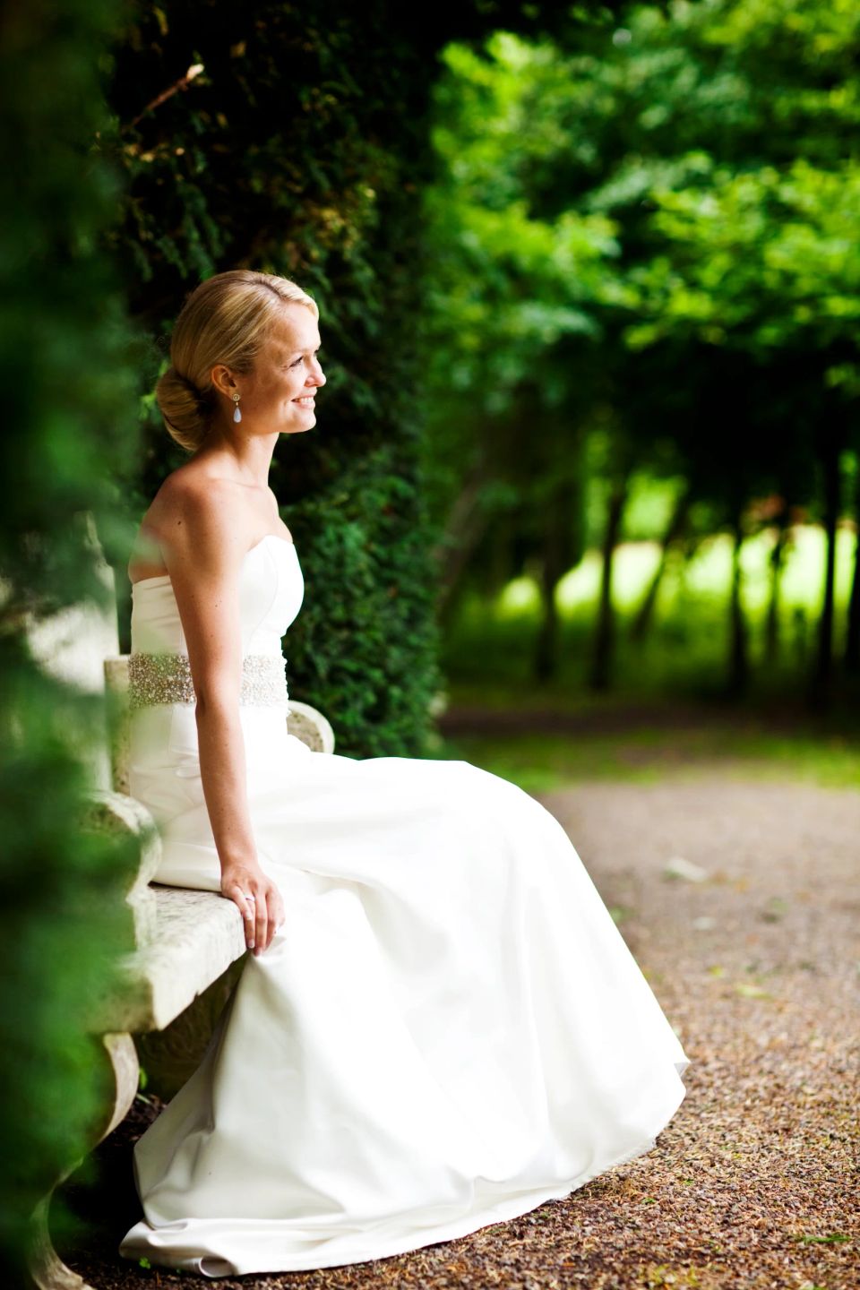 Brud i hvid kjole sidder smilende på stenbænk i grøn have. Et autentisk bryllupsfoto i naturligt lys.