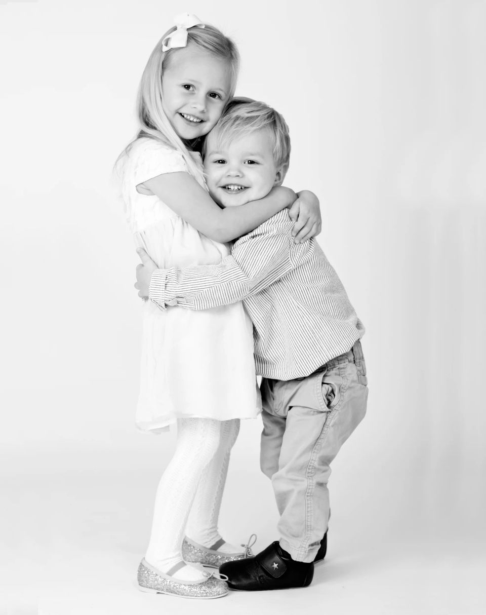 Børn i Virum: Sort/hvidt studieportræt af to smilende søskende, der krammer hinanden tæt og kærligt.