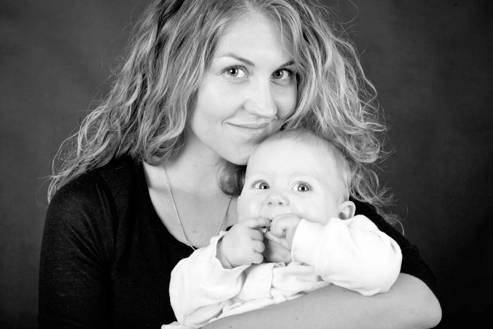 Sort/hvidt familiefoto af mor der smilende holder baby med store øjne og fingre ved munden i blødt lys.