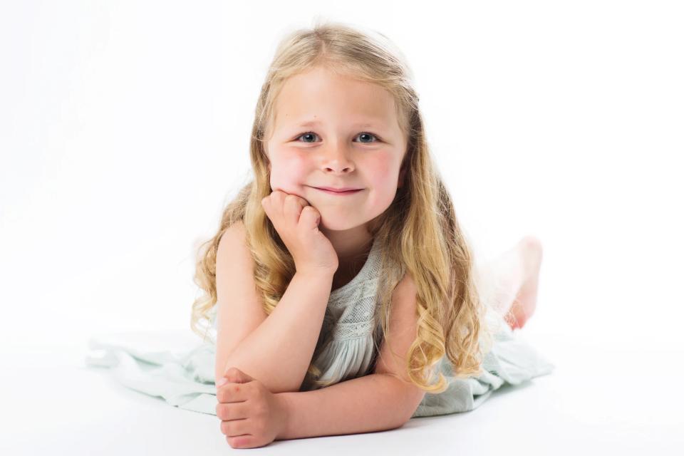 Børnefotografering af smilende, lyshåret pige i lysegrøn kjole, der ligger afslappet på maven mod hvid baggrund.