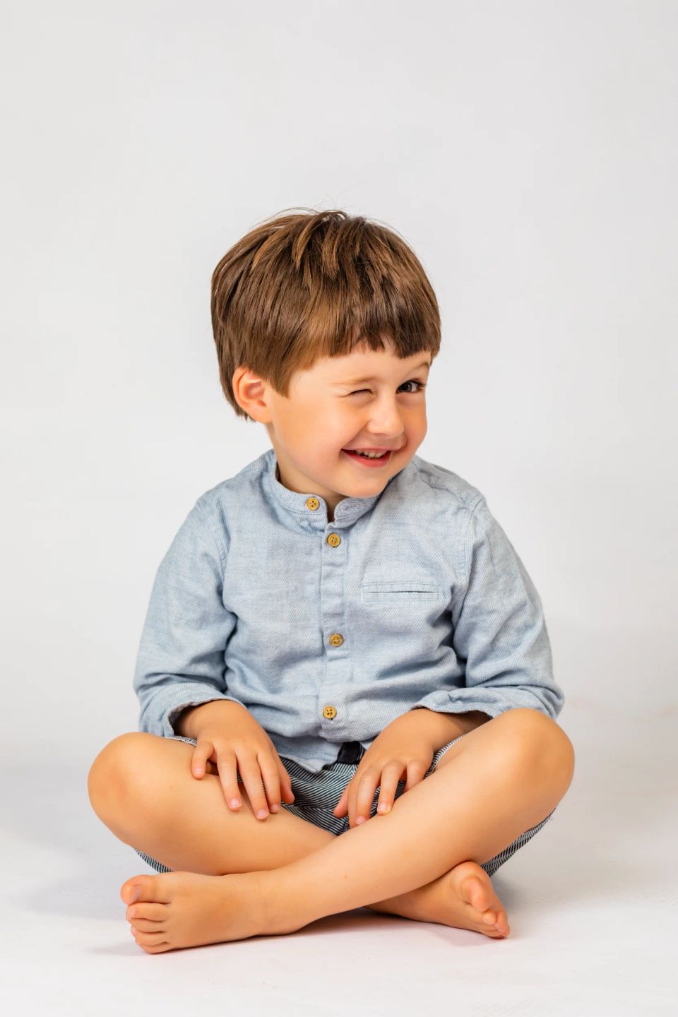 Glad dreng med brunt hår og blå skjorte, med et legende glimt i øjet, sidder og smiler. Et skønt børnefoto.