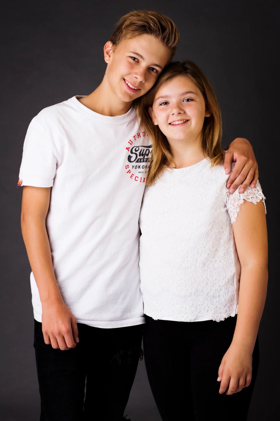 Familiefoto af smilende søskende, en dreng og en pige, i hvide trøjer mod en mørk baggrund i naturligt lys.