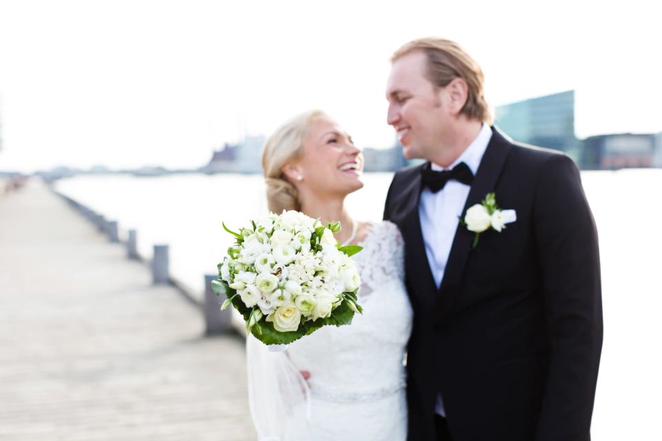 Bryllupsfoto af smilende brudepar ved vandet, med bruden holdende en hvid buket i blødt dagslys.