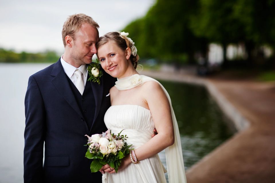 Bryllupsfoto af smilende brudepar i favn udendørs med sø og grønne træer i blød baggrund. Bruden holder buket.