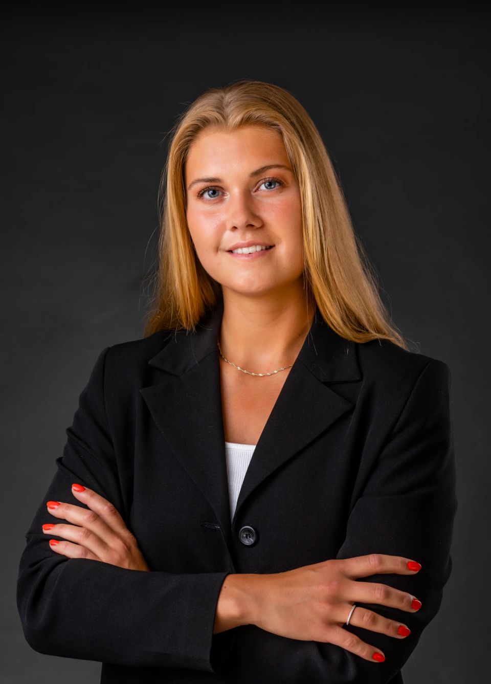 Professionelt portrætfoto af ung, blond kvinde smilende med armene krydset, iført sort blazer på mørk baggrund.