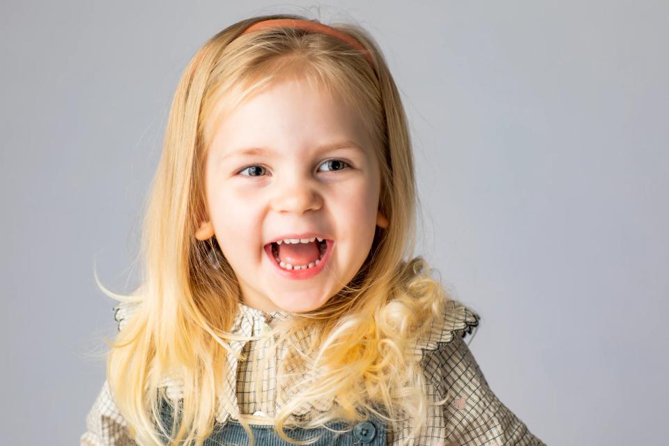 Glad, blond pige med åben mund og smilende øjne, iført ternet bluse og lyseblå selekjole. Familie fotografering.