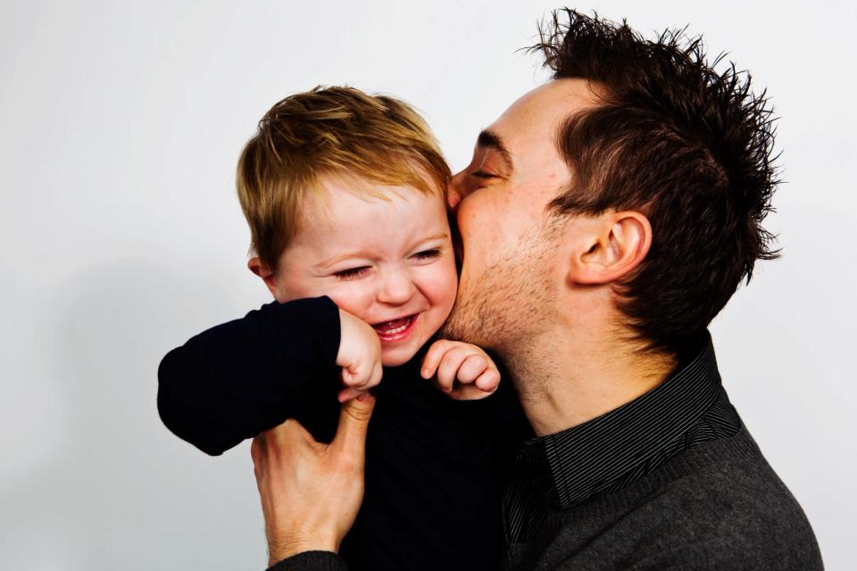 Far i mørk trøje kysser sin glade baby på kinden. Barnet griner, holdt af sin fars hånd. Smukt familiefotografi.