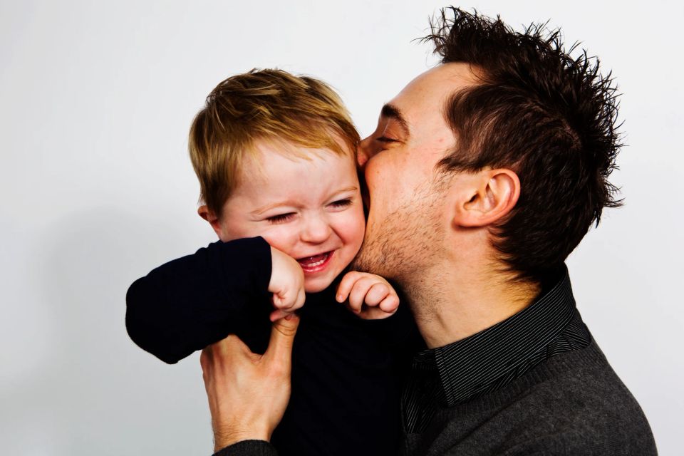 Far i mørk trøje kysser sin glade baby på kinden. Barnet griner, holdt af sin fars hånd. Smukt familiefotografi.