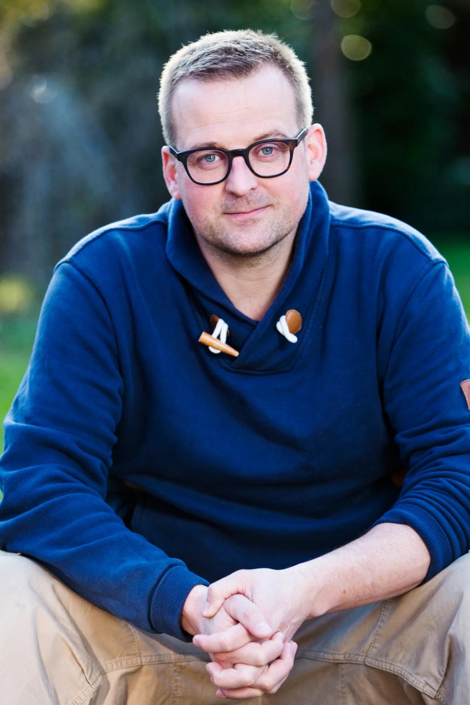 Portrætfoto af mand med briller i Virum, iført mørkeblå trøje, med let smil og foldede hænder.