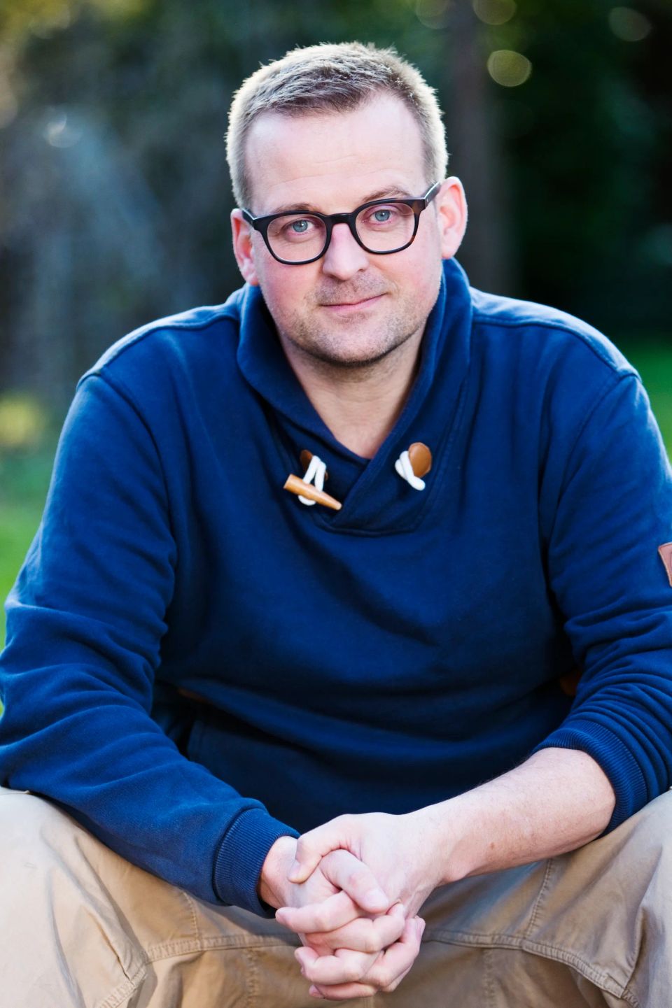Portrætfoto af mand med briller i Virum, iført mørkeblå trøje, med let smil og foldede hænder.