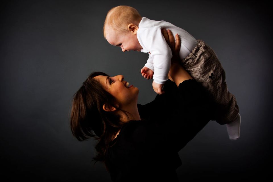 Glad mor løfter sin baby op i luften, set i profil, mod en mørk baggrund. Babyfotografering med varme, bløde farver.