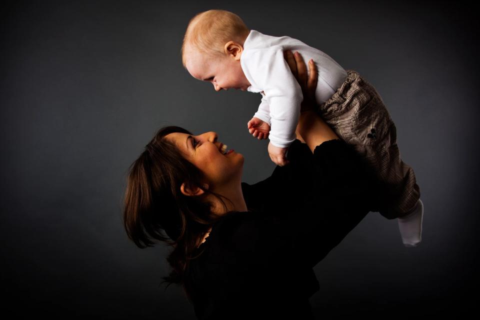 Glad mor løfter sin baby op i luften, set i profil, mod en mørk baggrund. Babyfotografering med varme, bløde farver.