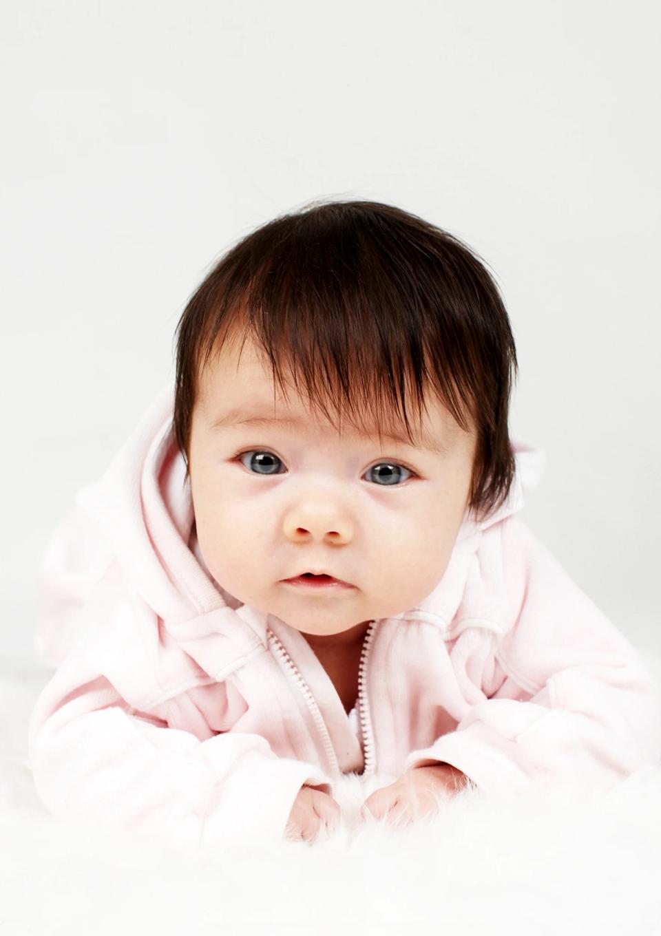 Nysgerrig baby i rosa trøje med mørkt hår og store øjne, liggende på hvidt tæppe. Et rørende baby portrætfoto.