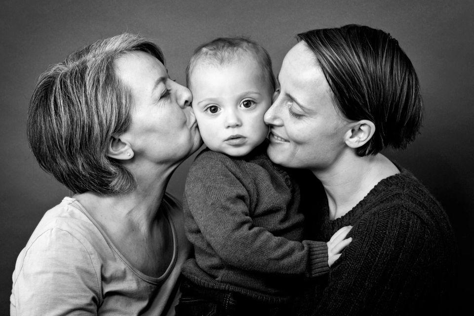 Sort/hvid familiefoto af mor og mormor der kysser en baby på kinderne, hvilket skaber et hjertevarmt øjeblik.