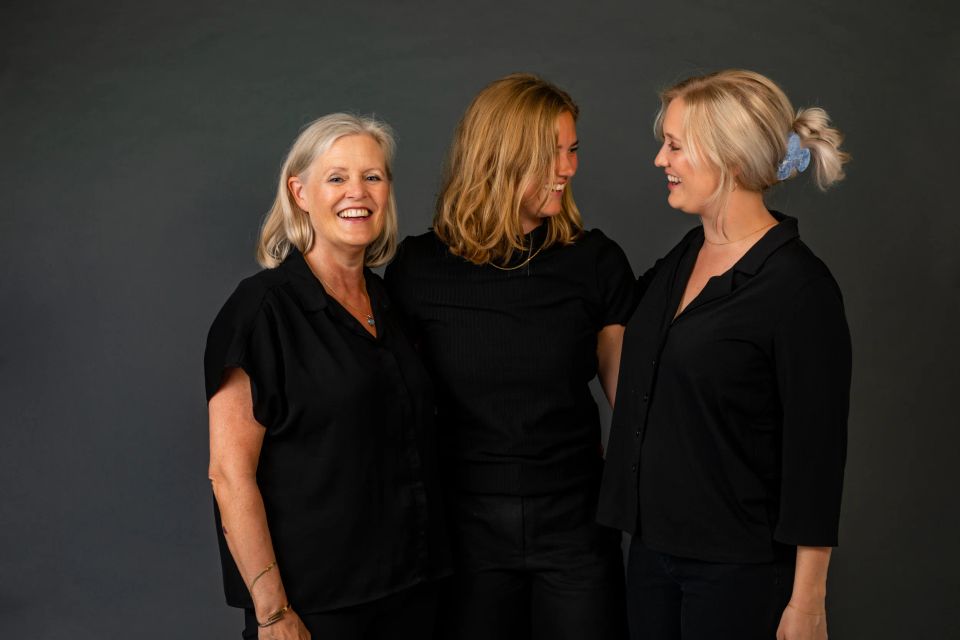 Familiefotografering med tre kvinder i sorte bluser, der smiler og kigger på hinanden i blødt lys.