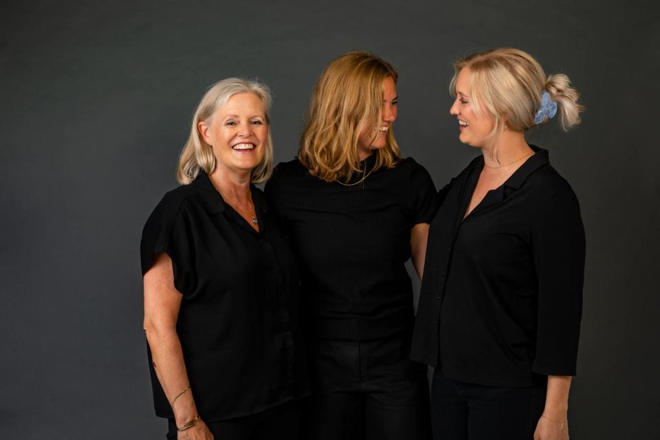 Familiefotografering med tre kvinder i sorte bluser, der smiler og kigger på hinanden i blødt lys.